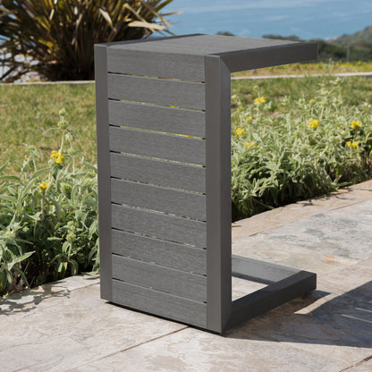 The Aluminum Outdoor C-Shaped Side Table for Patio, featuring horizontal slats and a modern gray finish, sits on a stone patio beside green plants, yellow flowers, grass, and an ocean view in the background.