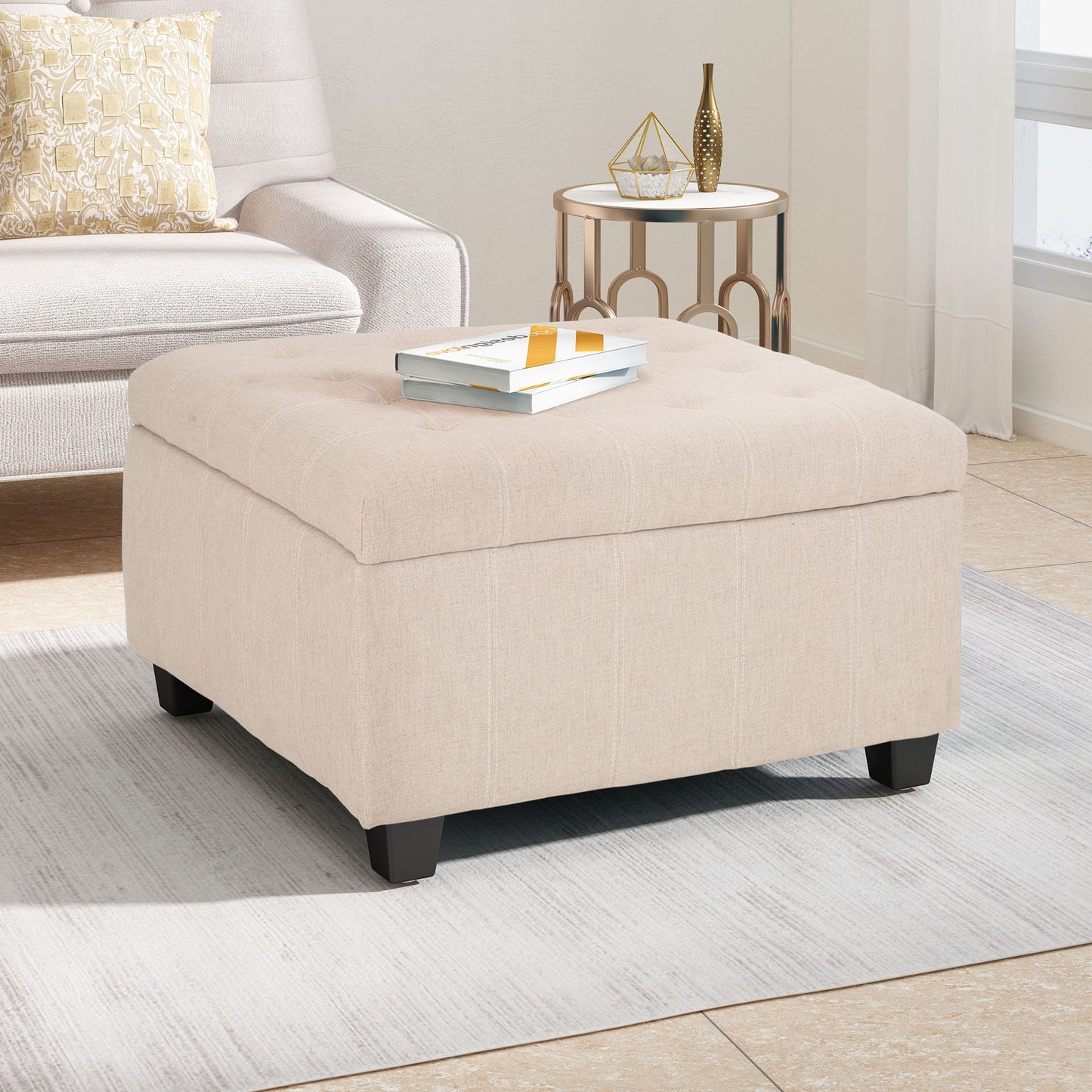 The Isabella Contemporary Ottoman With Storage sits on a light gray rug in a bright living room, next to a white armchair and a round side table with decor. Two books rest on top of the beige upholstered ottoman.