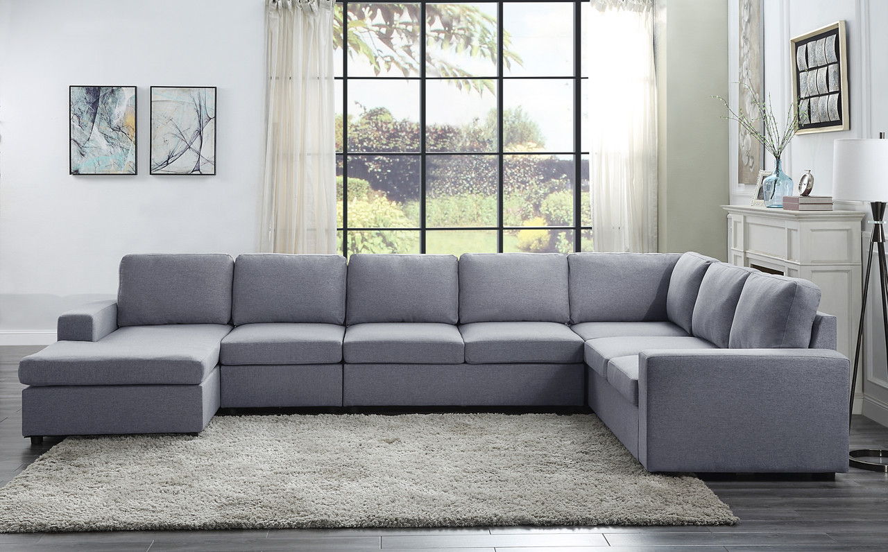 A modern living room features the Tifton - Linen 7 Seat Reversible Modular Sectional Sofa Chaise on a light rug with wooden floors. A big window reveals the garden, while two abstract paintings and a lamp add decorative touches.