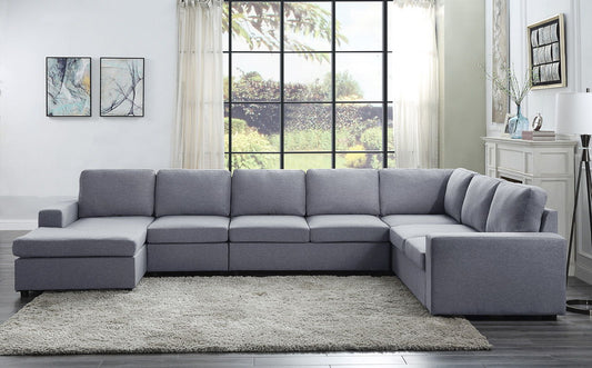 A modern living room features the Tifton - Linen 7 Seat Reversible Modular Sectional Sofa Chaise on a light rug with wooden floors. A big window reveals the garden, while two abstract paintings and a lamp add decorative touches.