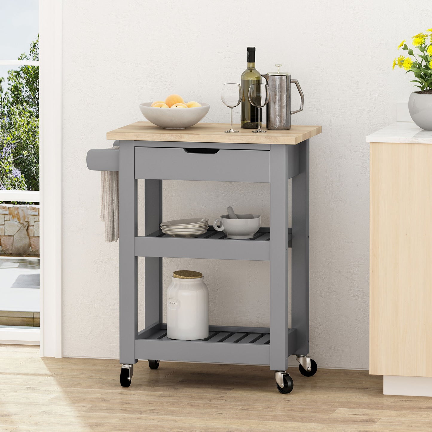 The Tattnall Kitchen Cart With Wheels, featuring a gray finish, wooden top, drawer, towel bar, and two shelves, holds kitchen essentials and is placed by a wall near a sunlit window.