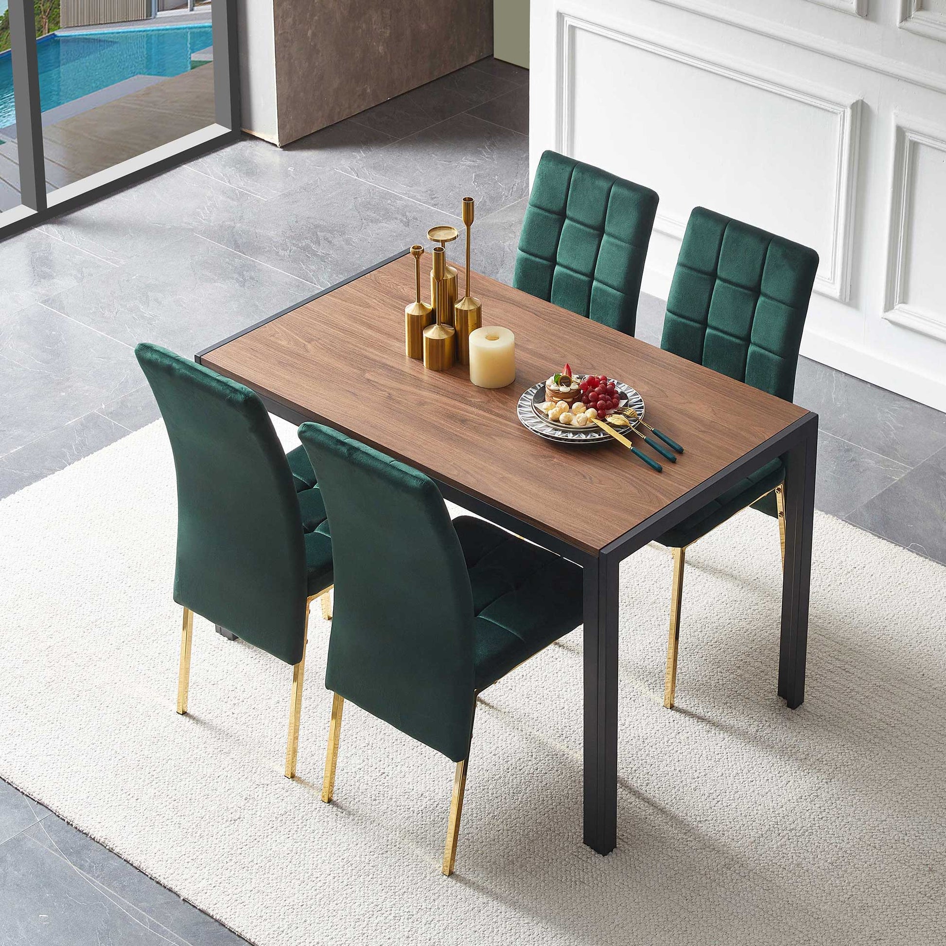 A 5-piece dining set with a creative rectangular table and four green velvet high-back chairs with gold legs, placed on a white rug. The table is topped with gold candlesticks, a candle, fruit, and cheese. A large window reveals a pool outside.