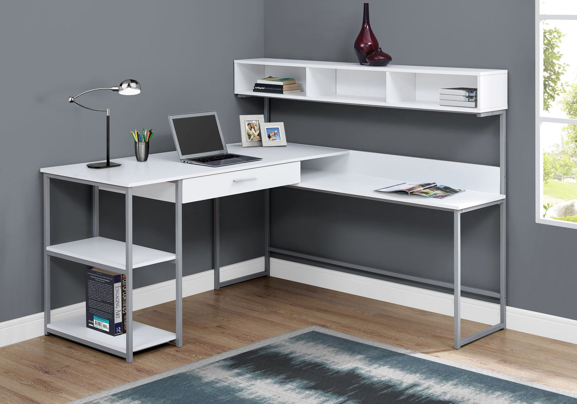 A contemporary white L-shaped computer desk with storage drawers holds a laptop, desk lamp, photos, and stationery. Books and decor rest on its shelves. Natural light enters through a window; the room features gray walls and a blue rug on wood flooring.