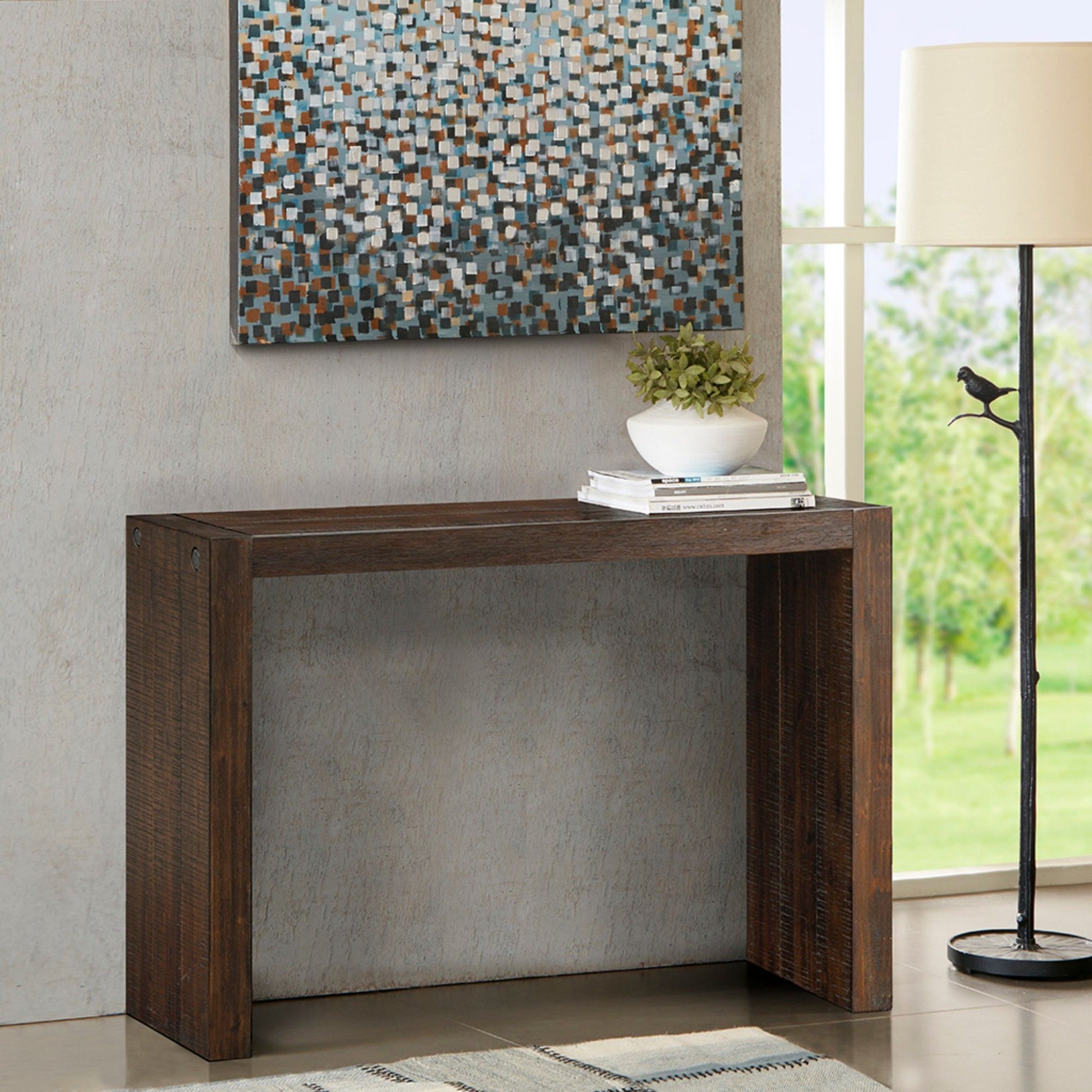 A modern entryway includes the Modern Design Console Table with a small plant and stacked books, a floor lamp, a patterned rug, and mosaic-style artwork displayed above the table.