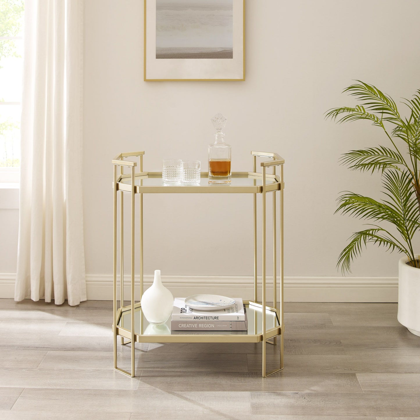 A Couch.com Modern Glam Mirror Top Accent Table in pale gold, with a decanter, glasses, a white vase, and books sits in a bright room with light wood floors, white curtains, a framed picture, and a green plant.