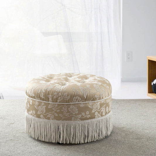 The Yolanda Round Upholstered Accent Ottoman, featuring a beige floral pattern and white fringe trim, sits on a light gray carpet in a bright, modern room with large windows and minimal furniture.