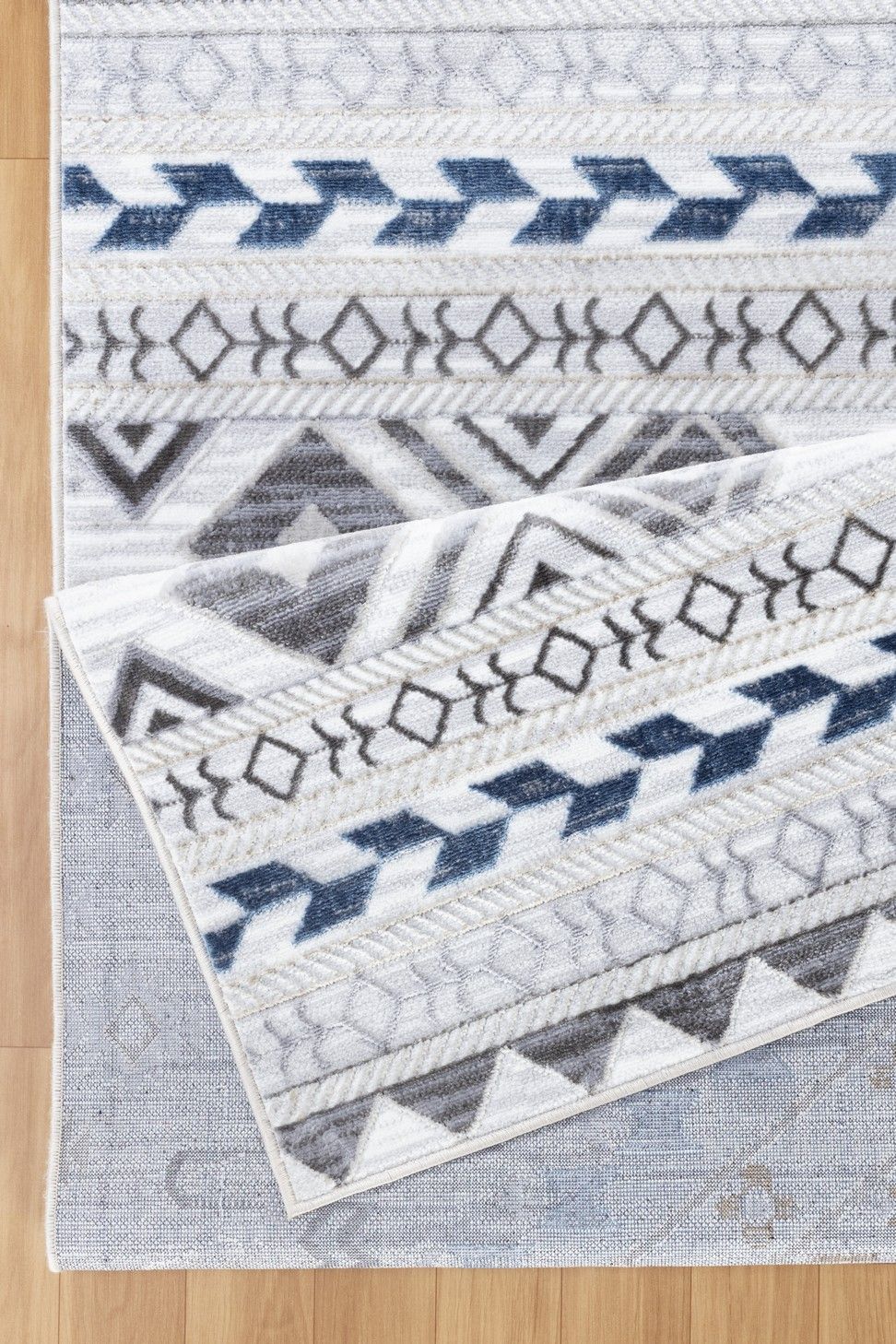 The Legacy - GC_CAM8001 Area Rug features a white and gray pattern with blue and black geometric designs, shown partially folded on a light wood floor to reveal a subtle blue and gray pattern beneath one corner.