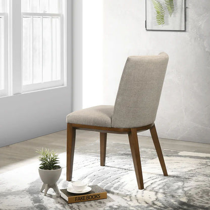 An Elmwood - Stylish Dining Chair (Set of 2) with beige upholstery and wooden legs is placed on a patterned rug near a window, next to stacked books labeled "FAKE BOOKS," a small plant, and a white cup and saucer.
