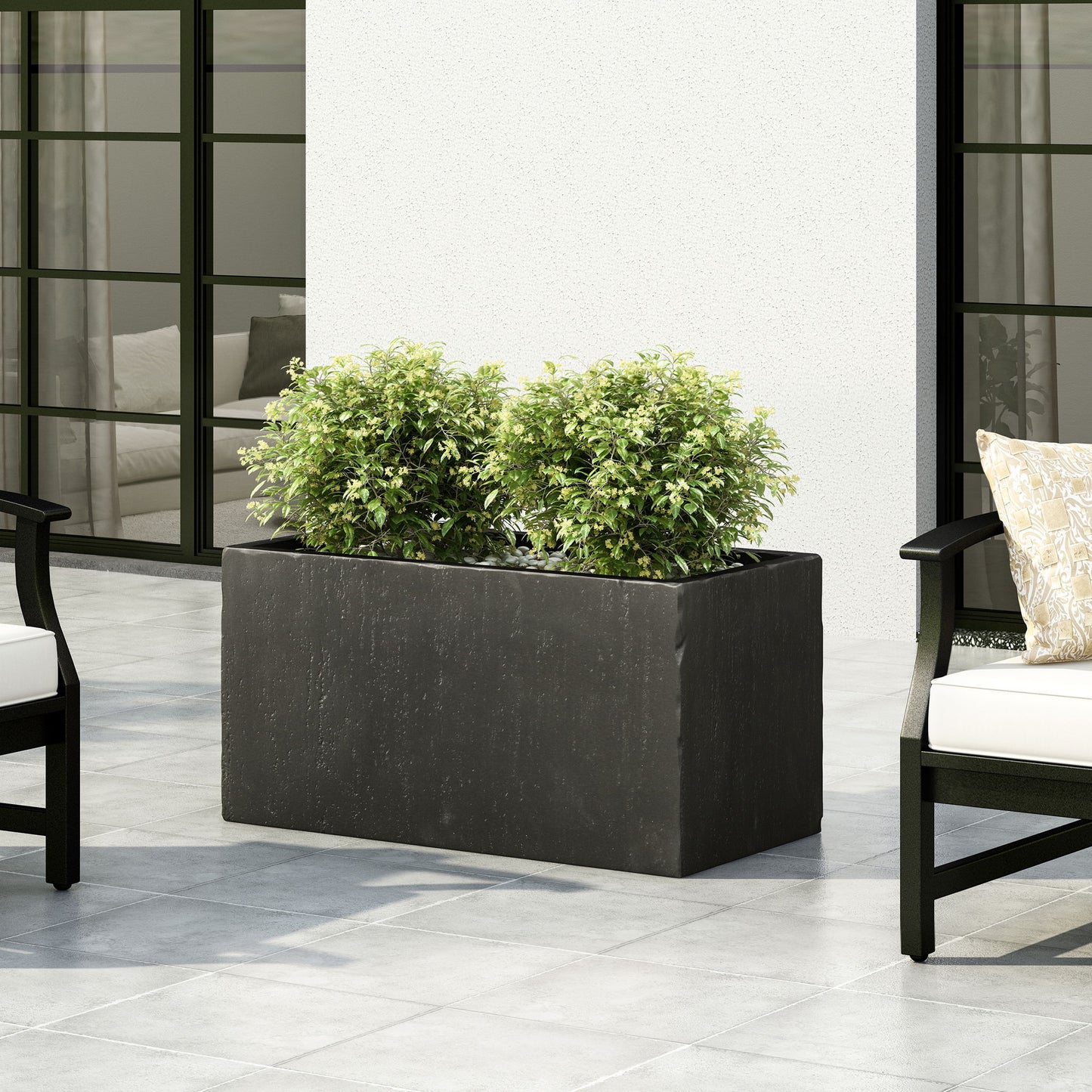 A Modern Outdoor Planter in black, holding two leafy green bushes, sits on a tiled patio between cushioned outdoor chairs, set against a white wall and large glass doors.
