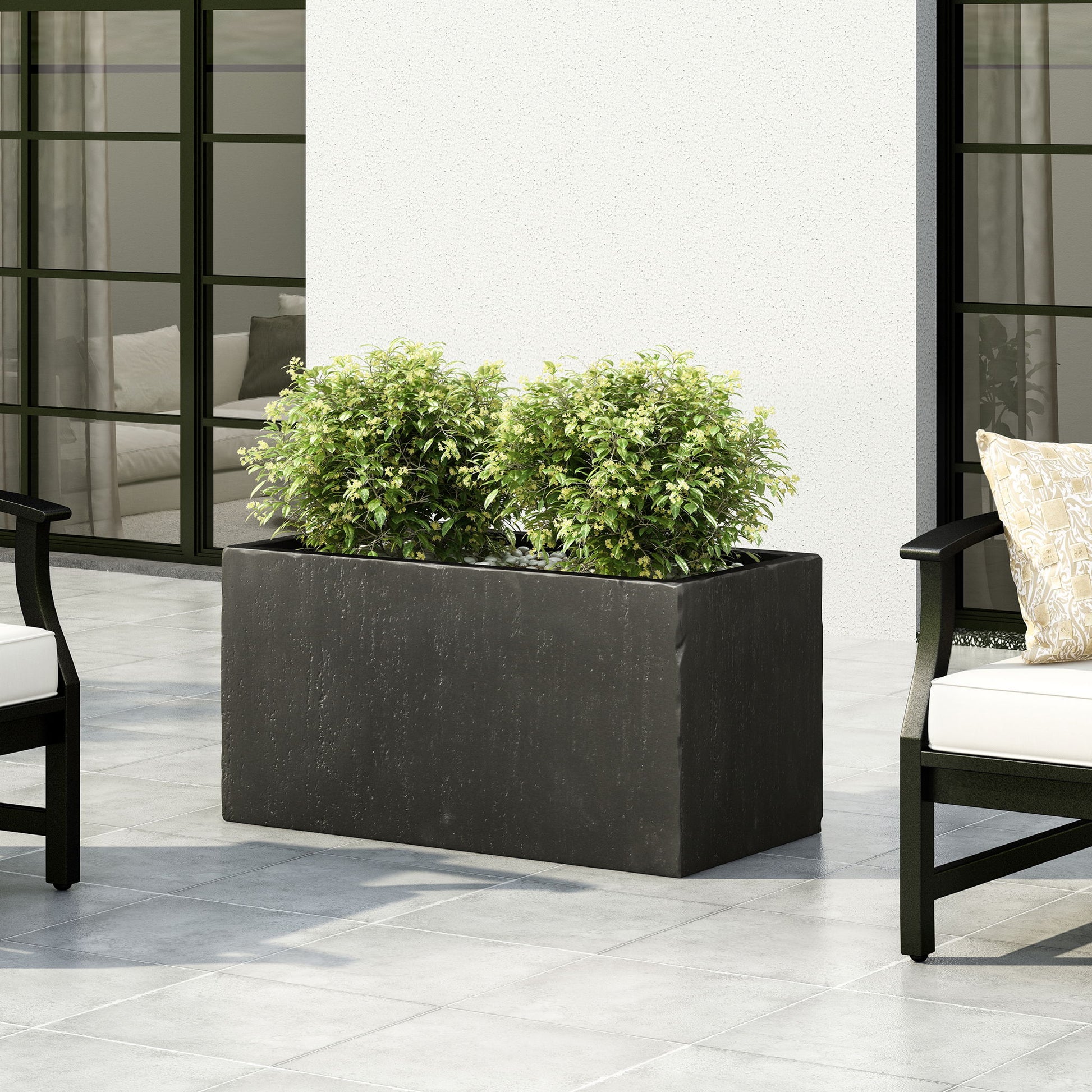 A Modern Outdoor Planter in black, holding two leafy green bushes, sits on a tiled patio between cushioned outdoor chairs, set against a white wall and large glass doors.
