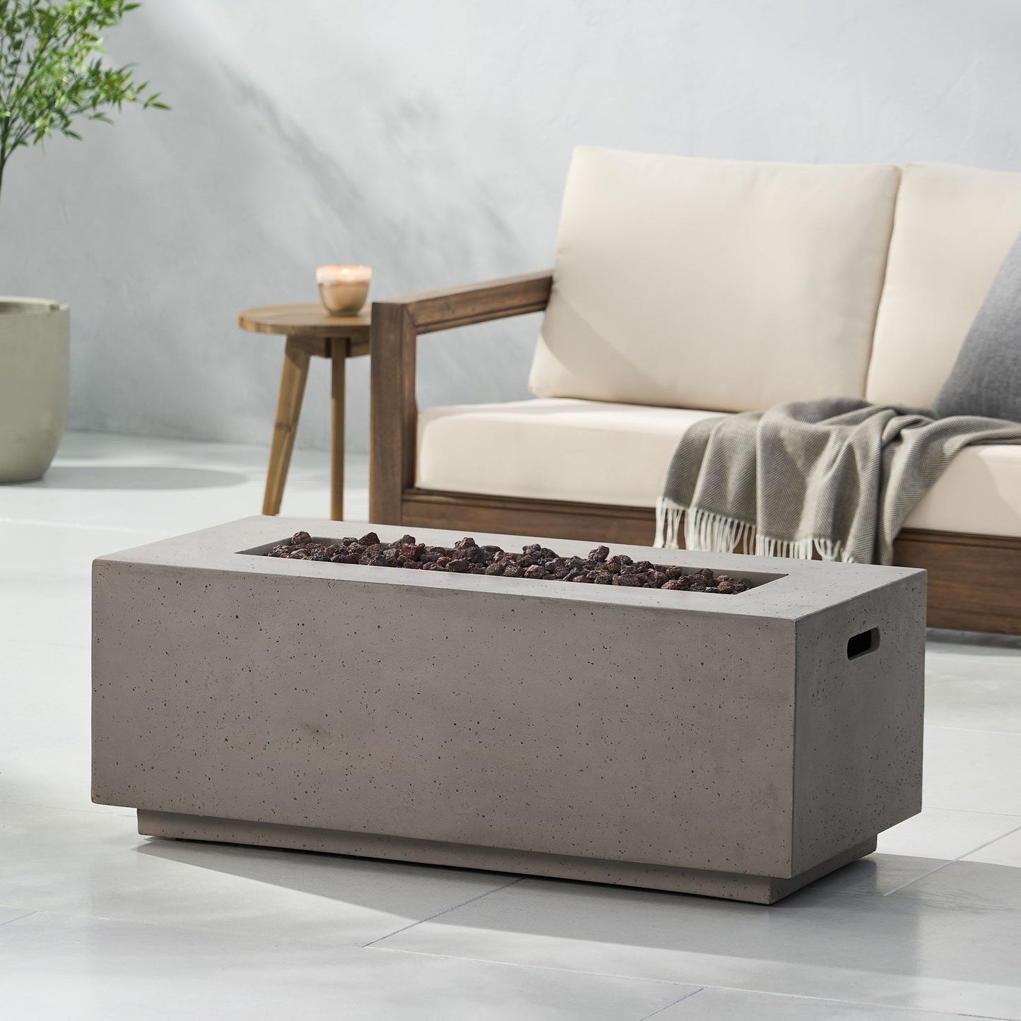 A modern patio features a beige cushioned sofa, a gray throw blanket, and the Contemporary Outdoor 40,000 Btu Propane Fire Pit filled with lava rocks. A potted plant and small candle rest nearby on the smooth, light-colored floor.