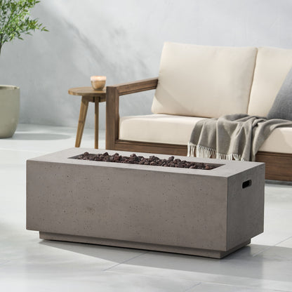 A modern patio features a beige cushioned sofa, a gray throw blanket, and the Contemporary Outdoor 40,000 Btu Propane Fire Pit filled with lava rocks. A potted plant and small candle rest nearby on the smooth, light-colored floor.