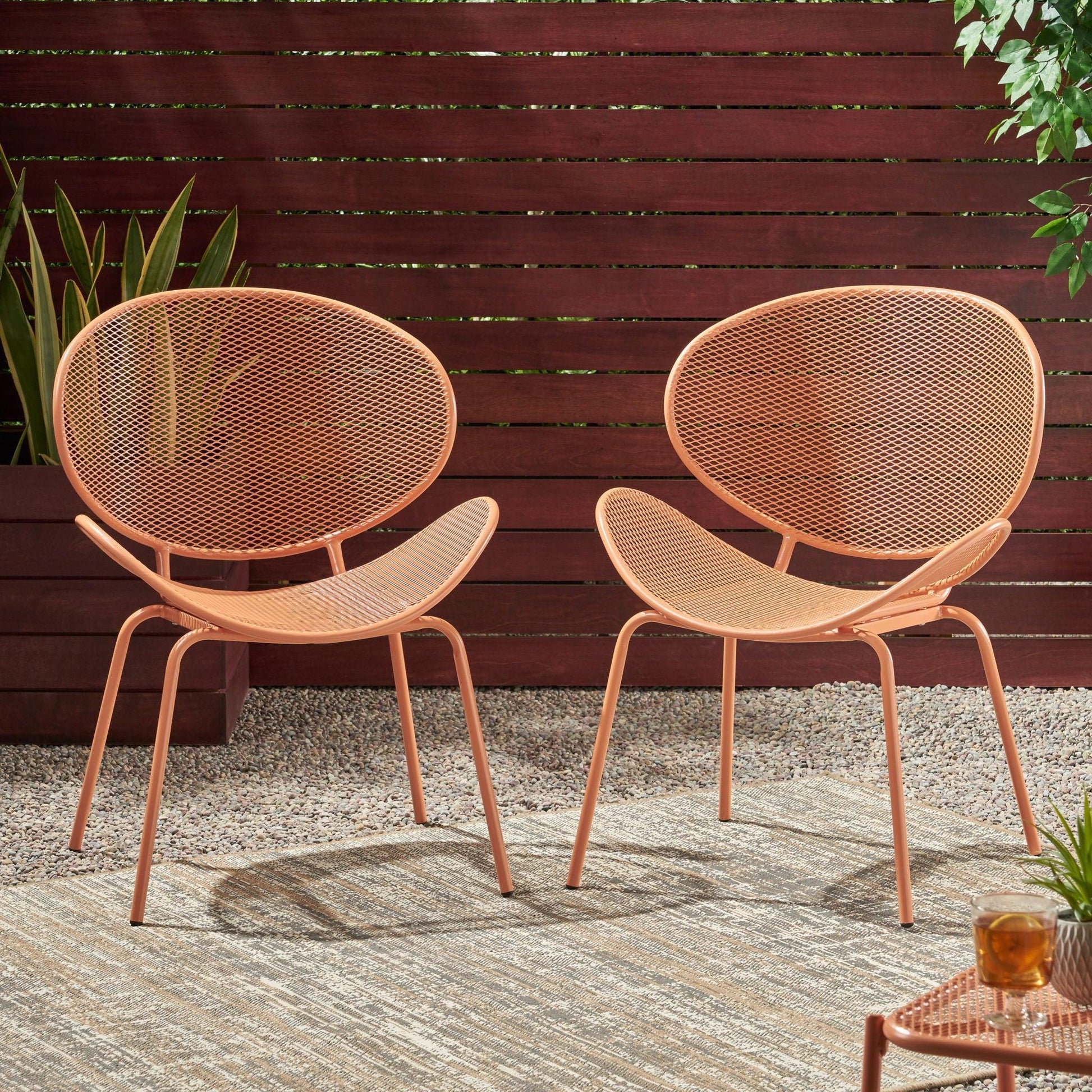 The Elba Contemporary Chair Set for Outdoor Relaxation features two modern orange metal chairs with mesh seats and backs, shown outdoors on a rug with gravel, a wooden fence, potted plants, and a small table with a drink nearby.