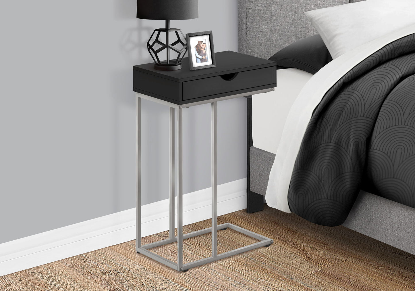 An Accent Table, C-Shaped Contemporary & Modern Design with a metal base, drawer, geometric lamp, and framed photo sits beside a bed with a black-and-white duvet in a bedroom with wood floors and gray walls.
