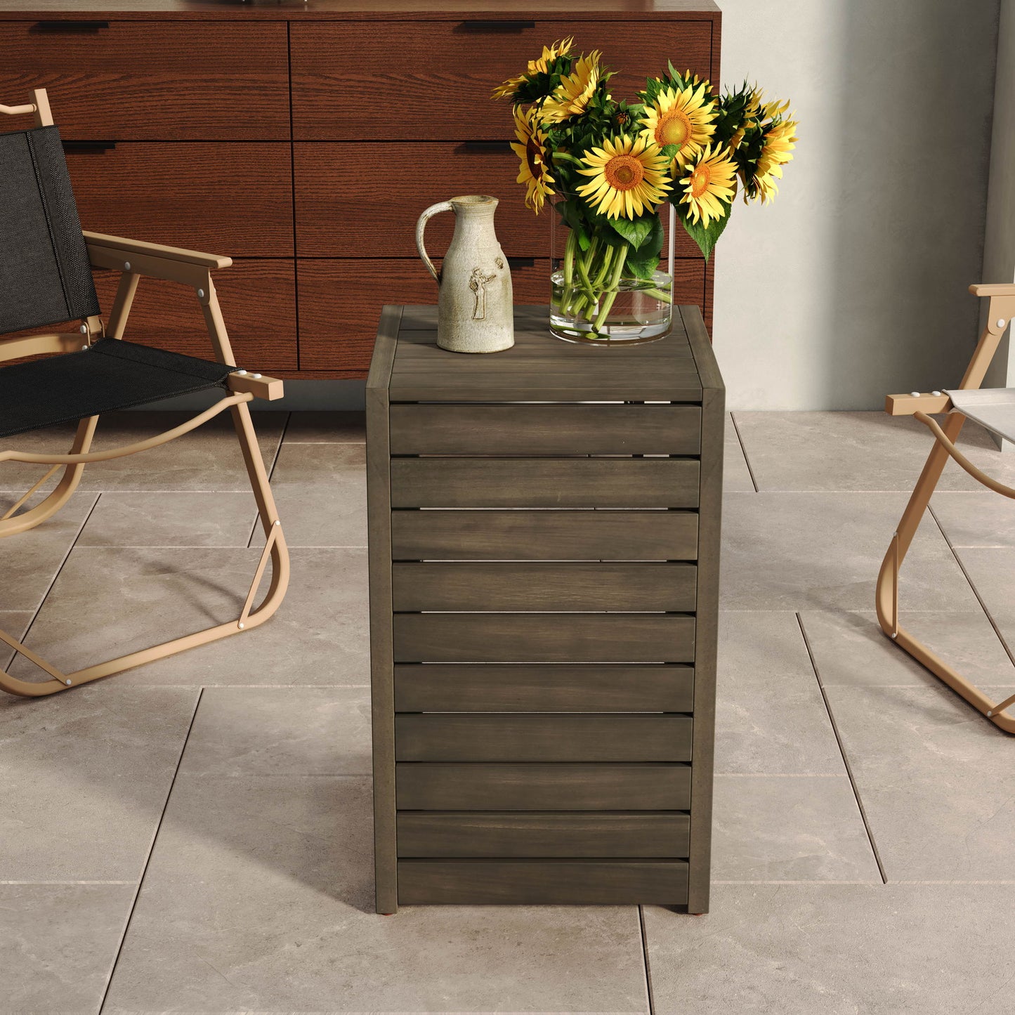 An Aluminum Outdoor C-Shaped Side Table for Patio holds a clear vase of sunflowers and a ceramic pitcher. Two folding chairs and a wooden dresser sit in the background on large beige tiles.