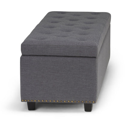 The Hamilton Upholstered Storage Ottoman features a rectangular gray design, tufted top, dark wood legs, and brass nailhead trim along the bottom edge.