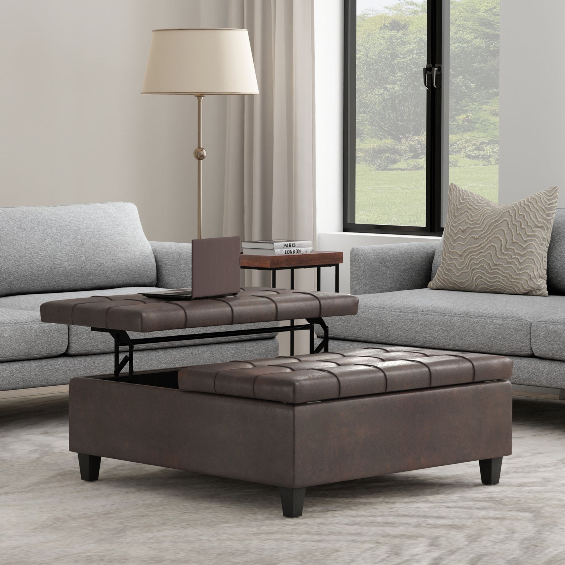 The Harrison Large Coffee Table Storage Ottoman, in brown and square-shaped with a lift-top tray, is placed between two gray sofas in the living room. A lamp and book are on a side table, and a large window fills the space with natural light.