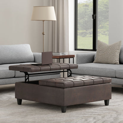 The Harrison Large Coffee Table Storage Ottoman, in brown and square-shaped with a lift-top tray, is placed between two gray sofas in the living room. A lamp and book are on a side table, and a large window fills the space with natural light.
