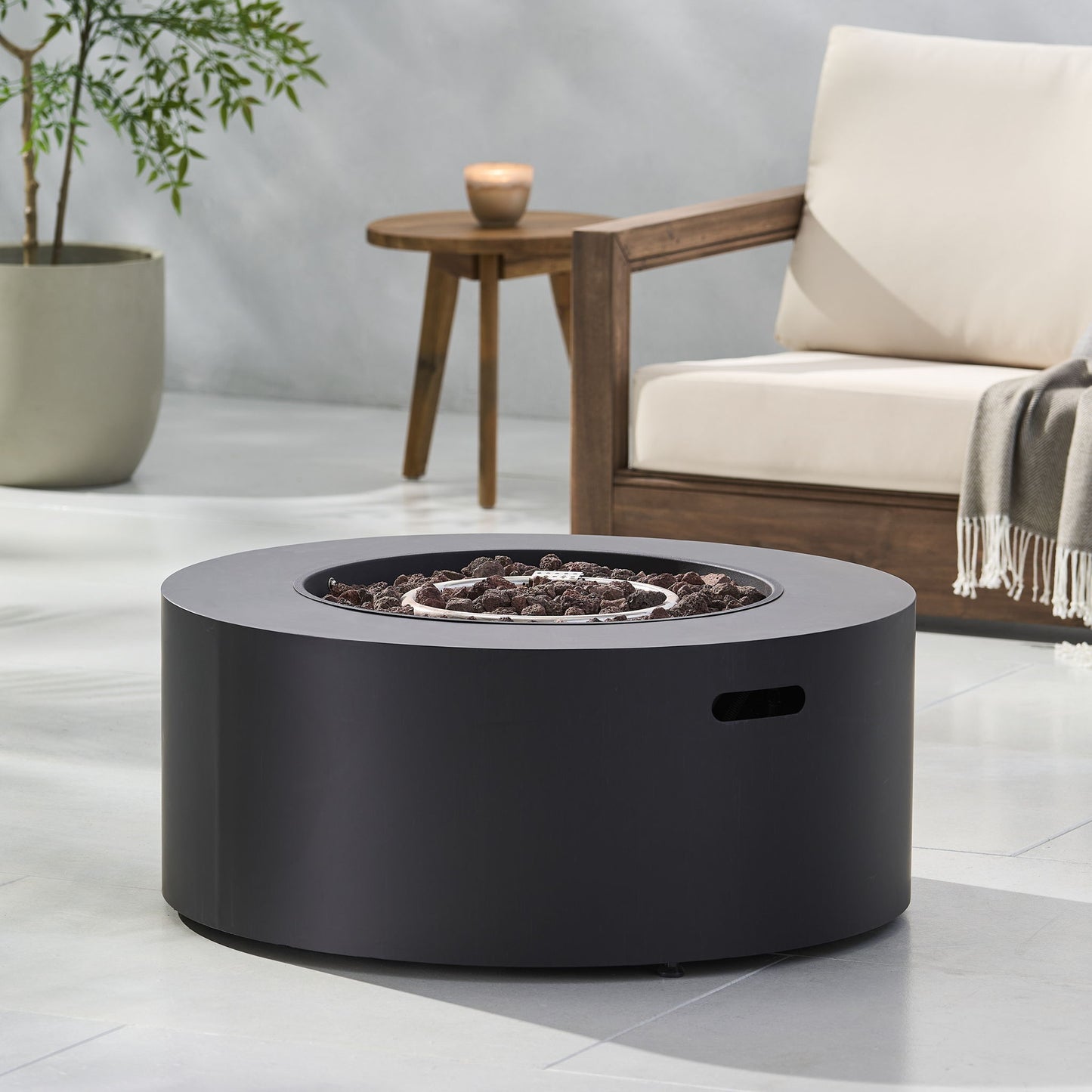 A Round Iron Fire Pit With 40,000 Btu featuring dark stones sits on a light patio beside a wooden chair with cream cushions, a throw blanket, and a small wooden side table with a candle. A potted plant is in the background.