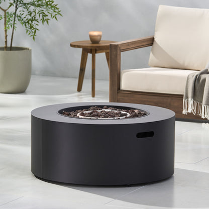 A Round Iron Fire Pit With 40,000 Btu featuring dark stones sits on a light patio beside a wooden chair with cream cushions, a throw blanket, and a small wooden side table with a candle. A potted plant is in the background.