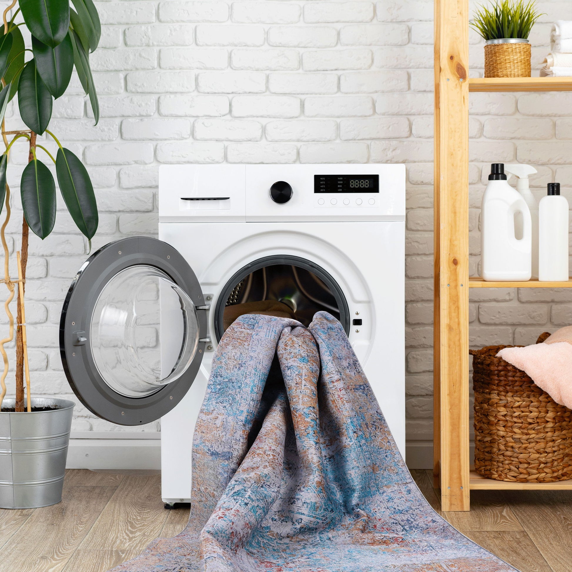 The Maval - Unique Washable Area Rug is partially placed inside a front-loading washing machine in a laundry room with a plant, shelf, detergent bottles, and wicker basket nearby.