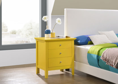 The Hammond 3 Drawer Nightstand in bright yellow stands beside a white bed with colorful bedding. Two vases of white flowers and a book rest on the nightstand, positioned near a large window that lets in natural light.