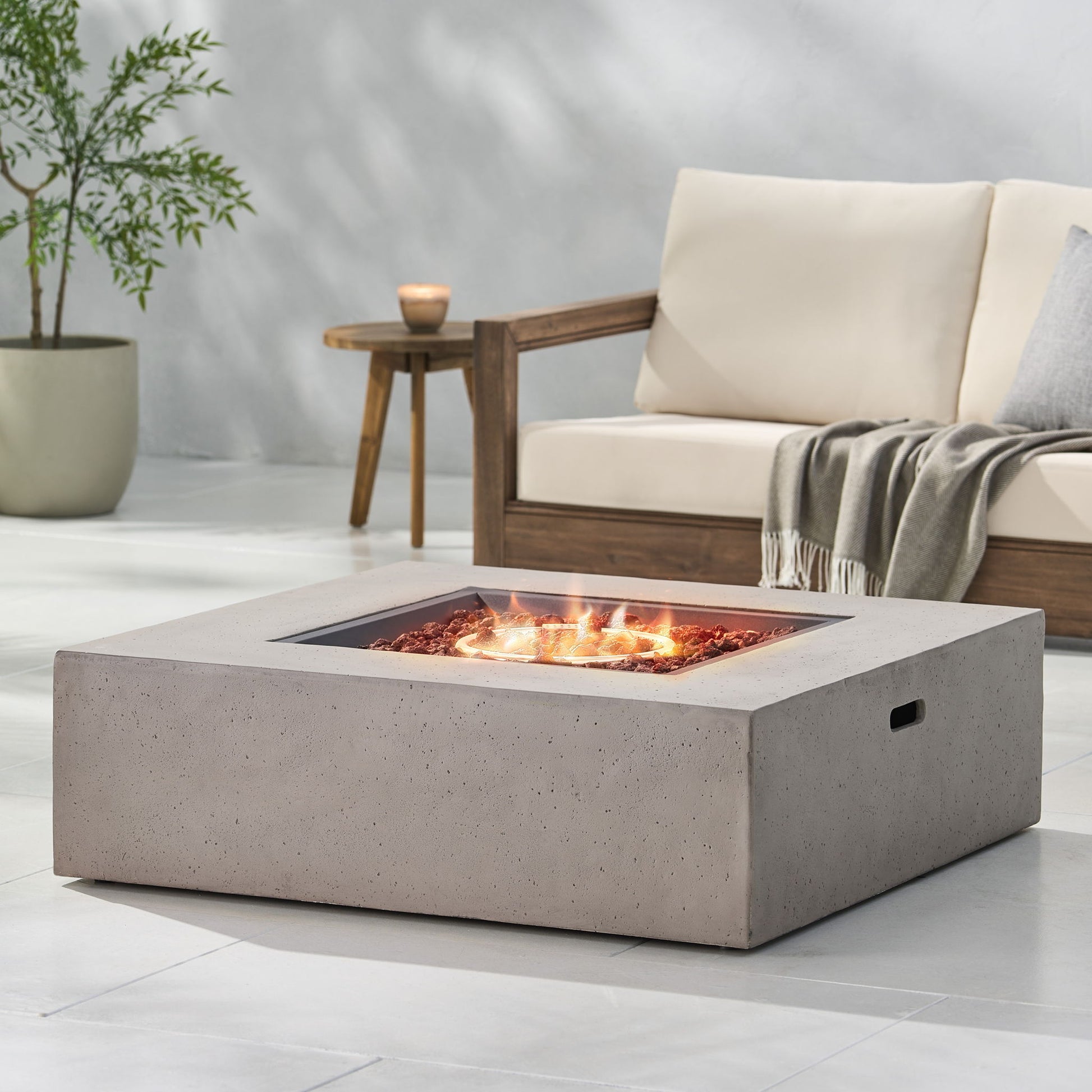 A modern patio features the Modern 50,000 Btu Square Fire Table With Propane Tank burning at the center. A wooden sofa with cream cushions, a gray throw, plus a potted plant and candle complete the inviting outdoor space.