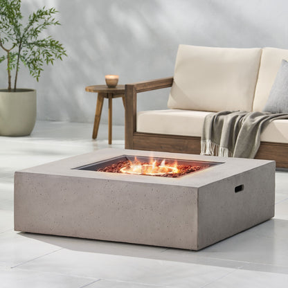A modern patio features the Modern 50,000 Btu Square Fire Table With Propane Tank burning at the center. A wooden sofa with cream cushions, a gray throw, plus a potted plant and candle complete the inviting outdoor space.