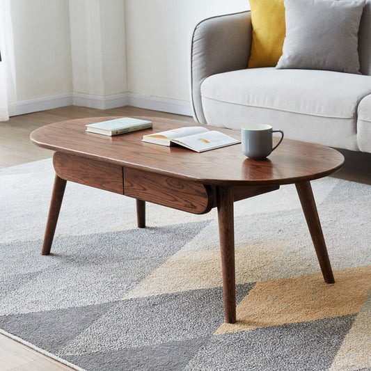 The Capsule Centre Low Table With Drawers Coffee Table with angled legs stands on a patterned rug, holding a cup and two books. Behind it, a light gray sofa is accented with a yellow cushion.