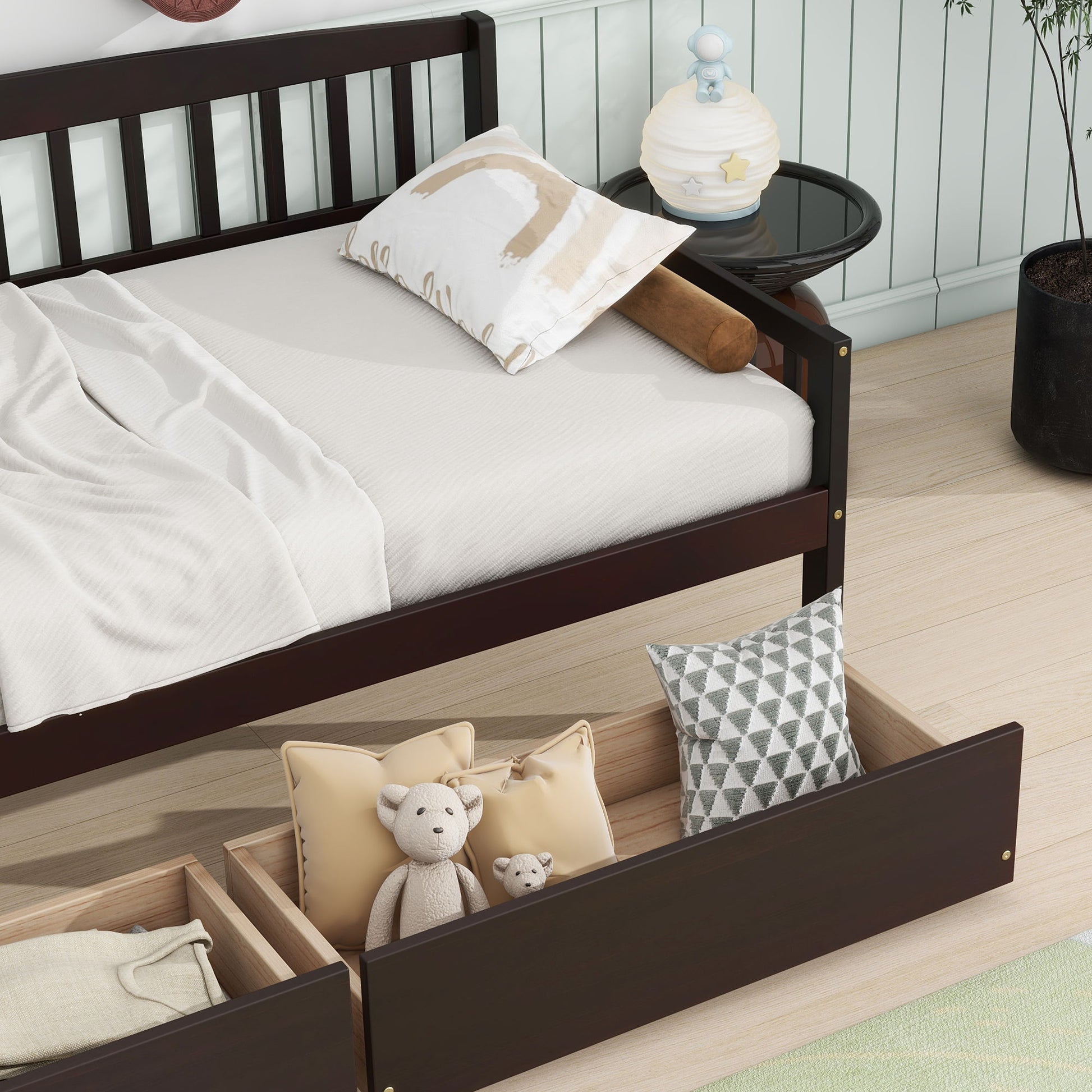 A Pine Wood Daybed with two storage drawers and a 10-slat platform sits on a light wood floor; drawers hold pillows and teddy bears. A round black side table and black plant pot are nearby, with light bedding on the dark wooden bed.
