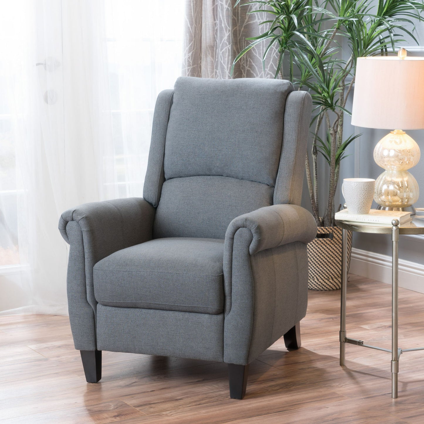 The Contemporary Push Back Recliner Chair in gray upholstery with rolled arms and dark legs sits on a wooden floor beside a round side table with a lamp, white mug, and plant, in a bright room with sheer curtains and a tall plant.