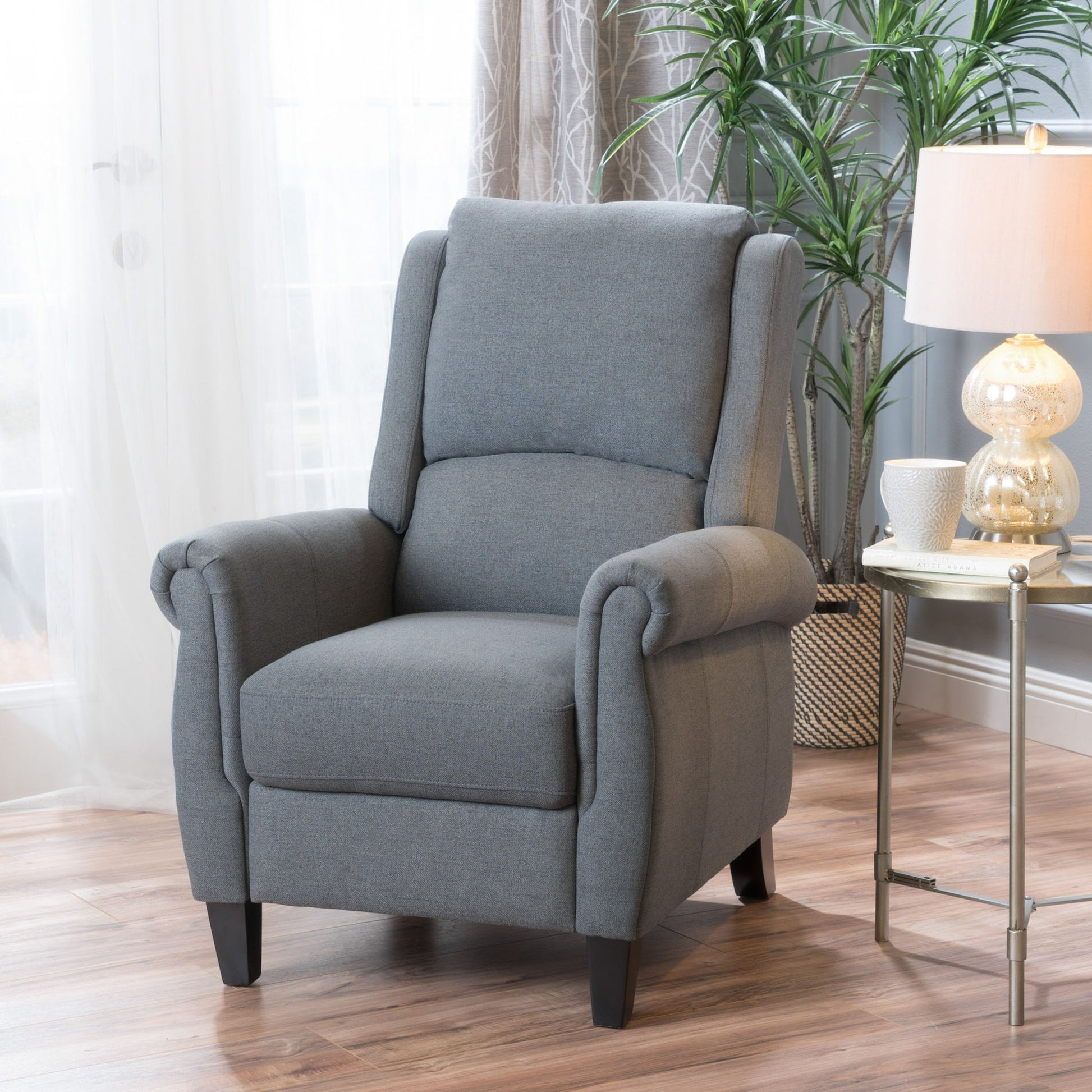The Contemporary Push Back Recliner Chair in gray upholstery with rolled arms and dark legs sits on a wooden floor beside a round side table with a lamp, white mug, and plant, in a bright room with sheer curtains and a tall plant.