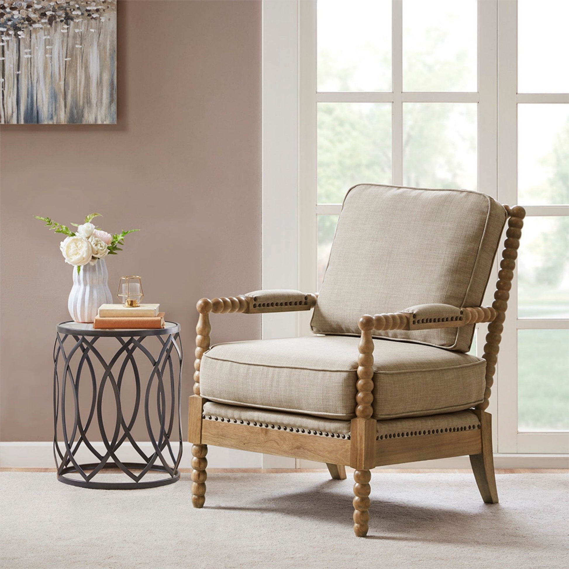 An Arm Chair With Open Back Spindle Design and padded armrests sits by a round metal side table with a vase of white flowers, book, and glass in a bright room with large windows and neutral walls.