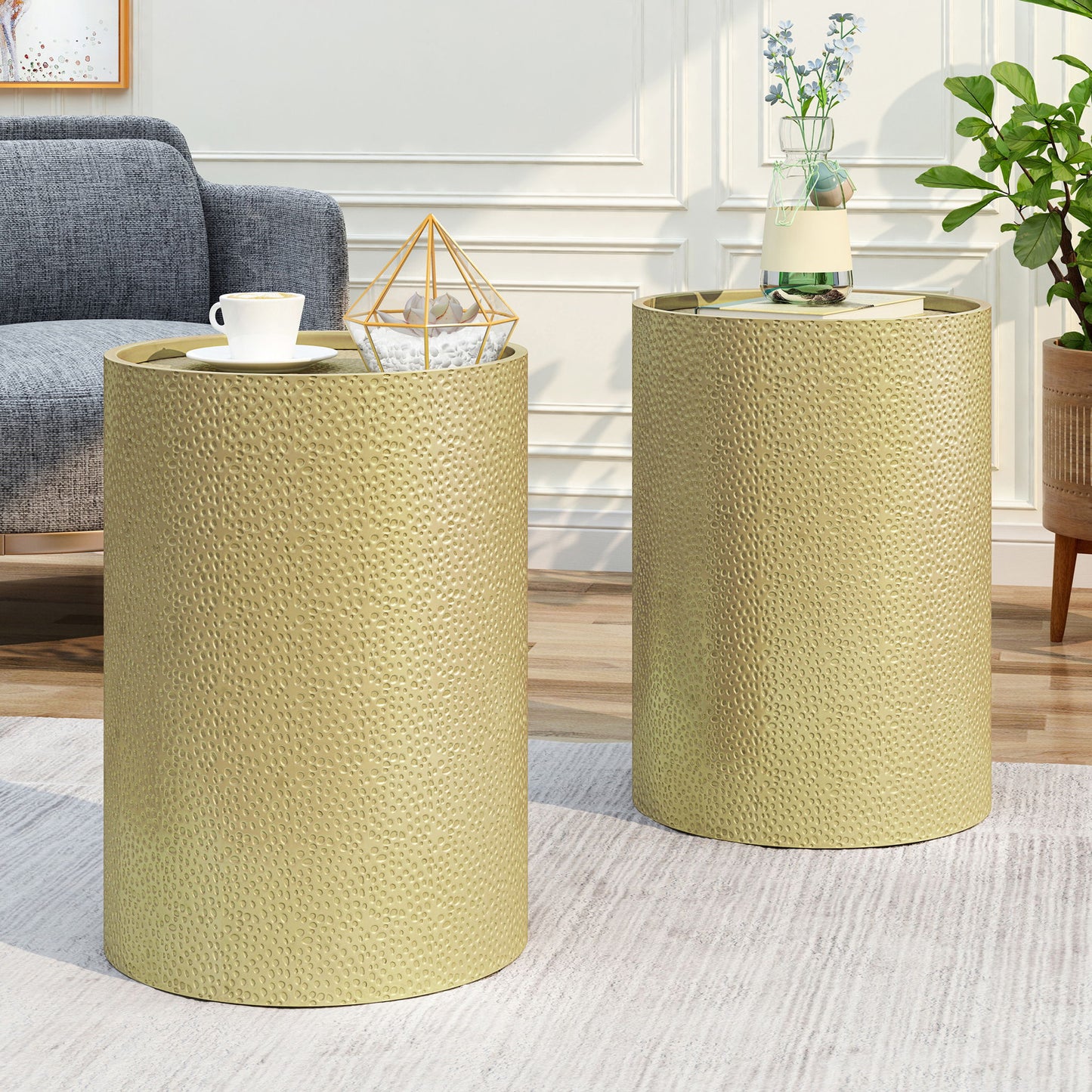 Two gold Modern Round Accent Tables With Hammered Surface sit in a modern living room—one holds a cup and geometric decor, the other a vase with flowers and a book. A gray sofa and plant are visible in the background.