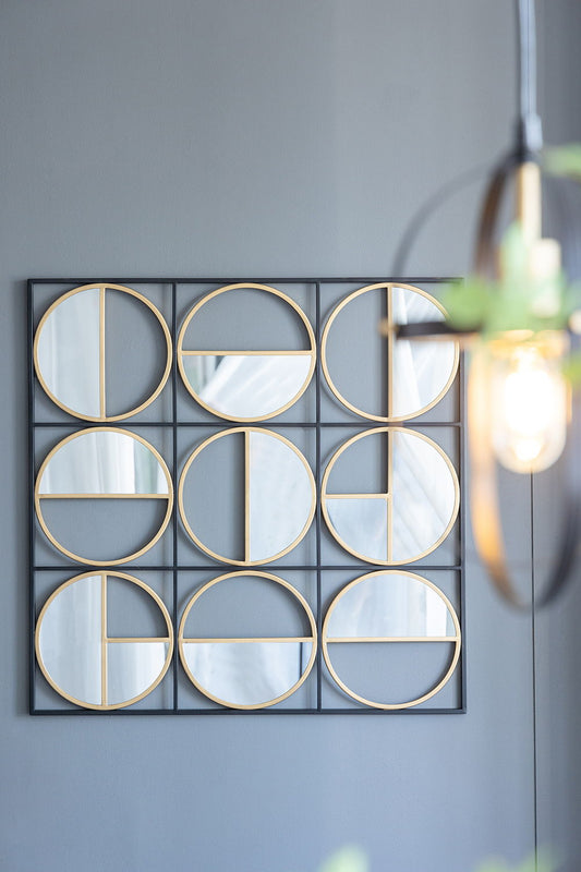 The Eclectic Styling Metal Beaded Wall Mirror with a contemporary geometric design and gold mirrored accents decorates a gray wall, while a modern light fixture and blurred leaves are visible in the foreground.