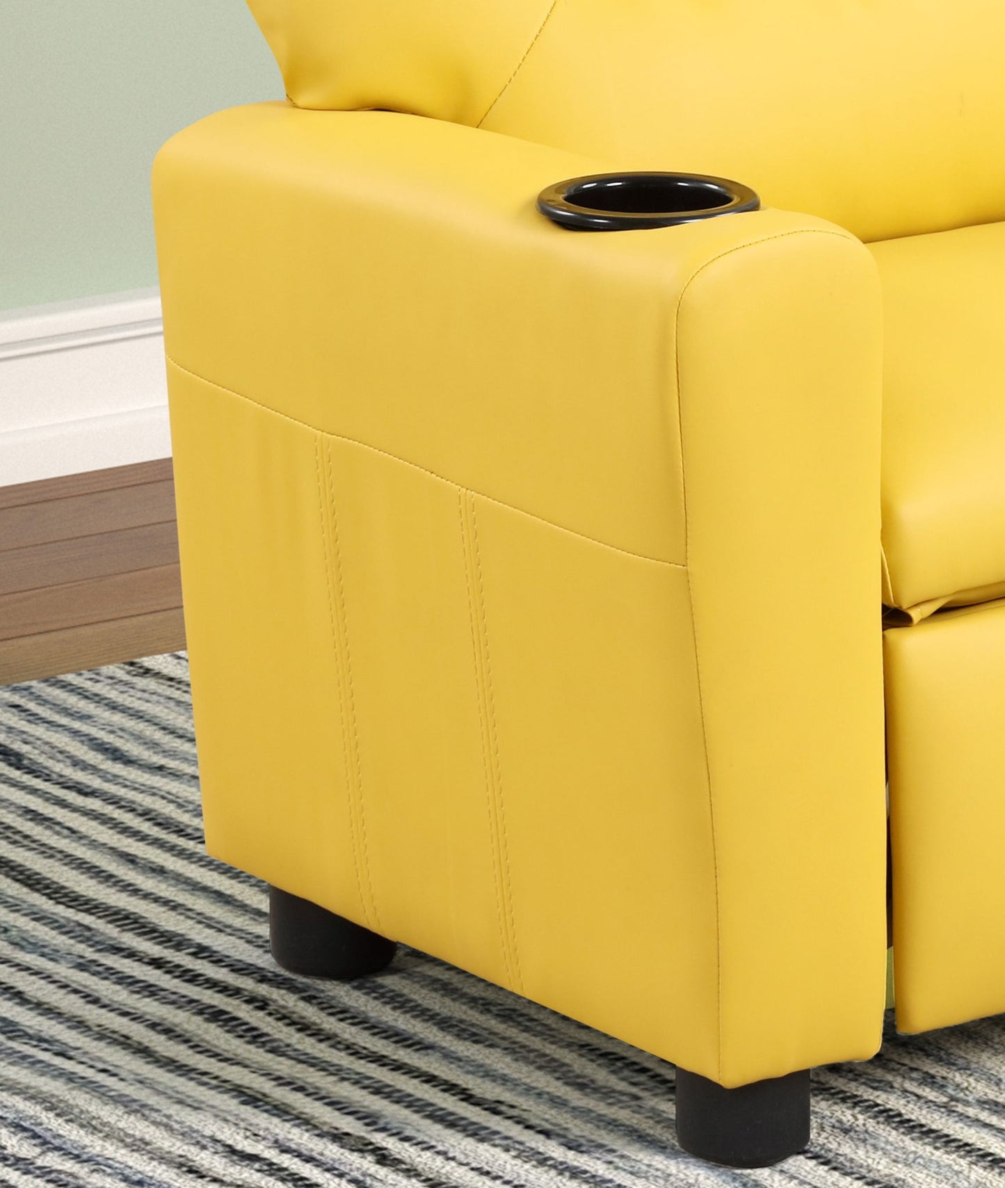 A close-up of the Marisa - PU Leather Kids Recliner Chair With Cupholder shows its yellow armrest and built-in black cupholder, set on a gray striped rug with a pale green wall and white baseboard in the background.