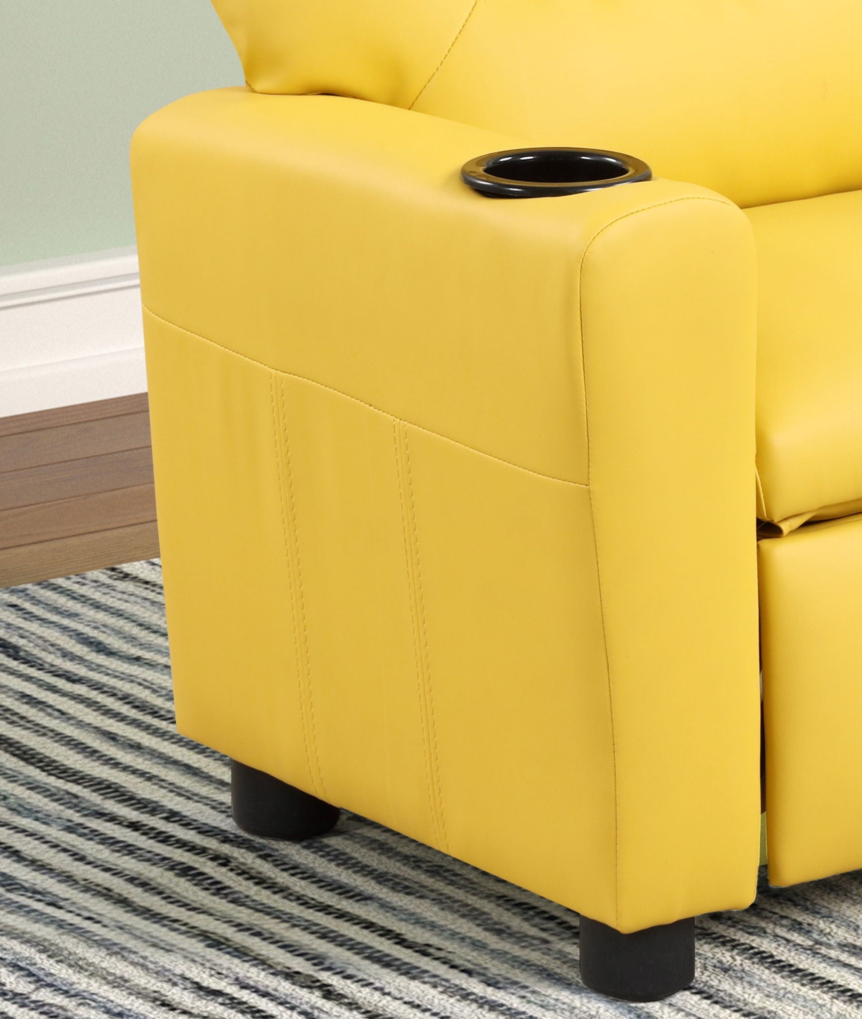 A close-up of the Marisa - PU Leather Kids Recliner Chair With Cupholder shows its yellow armrest and built-in black cupholder, set on a gray striped rug with a pale green wall and white baseboard in the background.