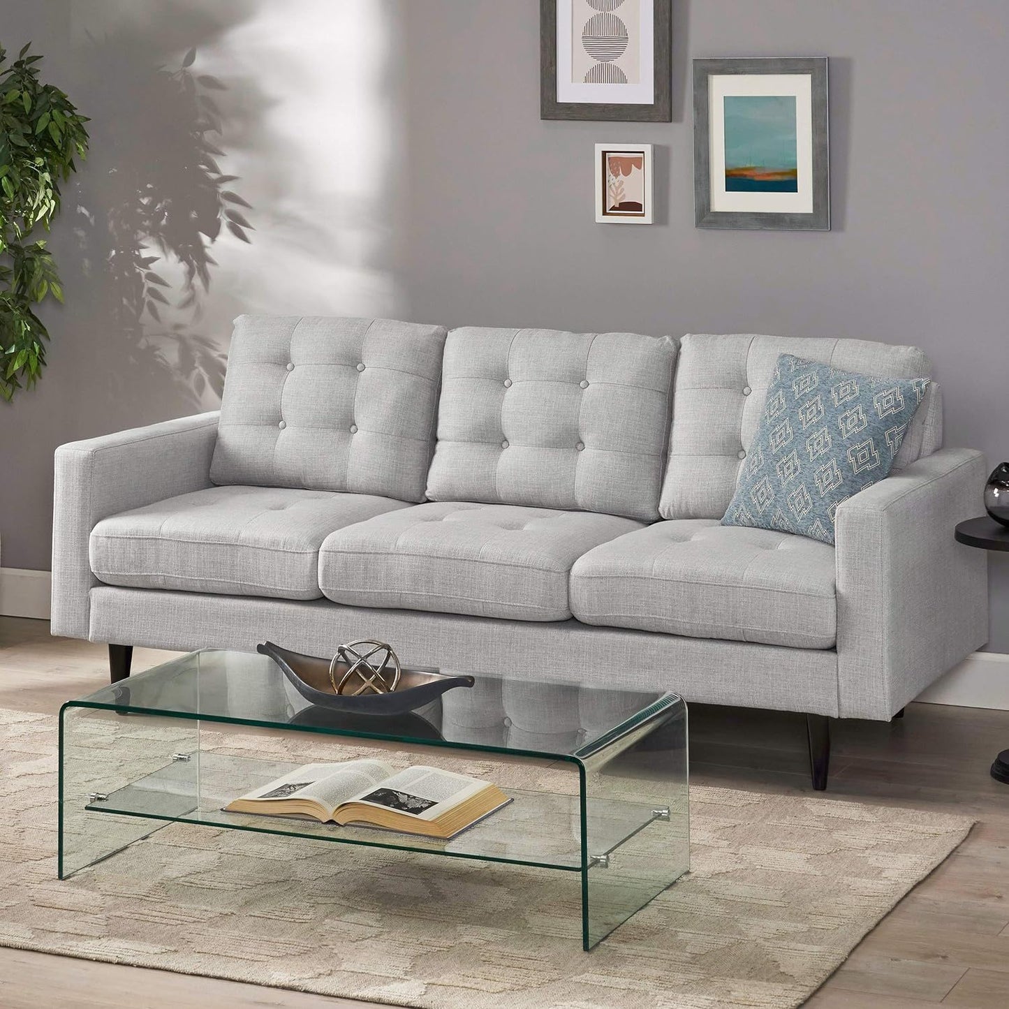 A modern living room with the Jenny Contemporary Tufted Fabric 3 Seater Sofa in light gray, a patterned blue pillow, glass coffee table with an open book and decor, beige rug, wall art, and a green potted plant in the corner.
