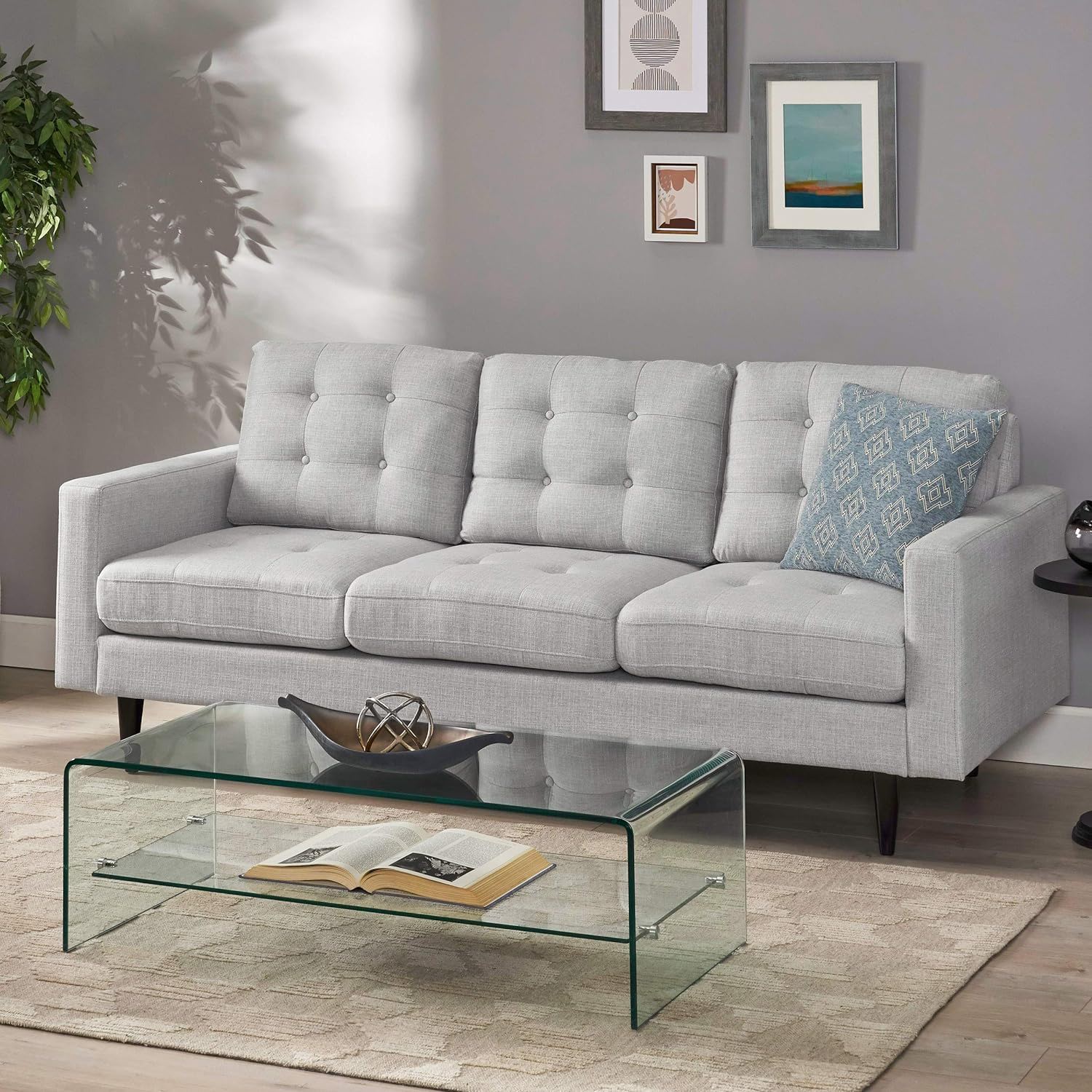 A modern living room with the Jenny Contemporary Tufted Fabric 3 Seater Sofa in light gray, a patterned blue pillow, glass coffee table with an open book and decor, beige rug, wall art, and a green potted plant in the corner.