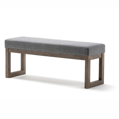 The Milltown Upholstered Ottoman Bench features a cushioned gray fabric seat and a wooden frame with open rectangular legs at each end, offering a simple, minimalist design.