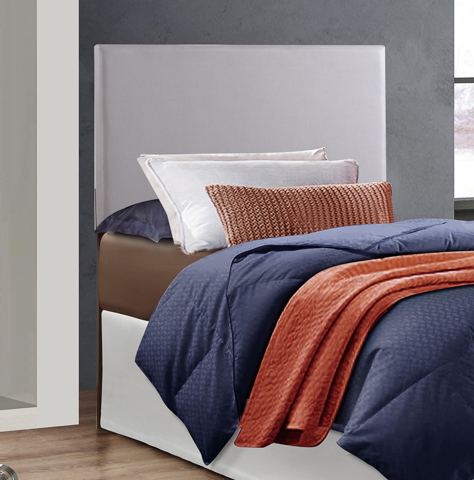 A neatly made bed features the Nova - Headboard in light gray upholstery, paired with a navy blue comforter, white and gray pillows, and a rust-colored throw blanket and pillow, set against a gray wall by the window.