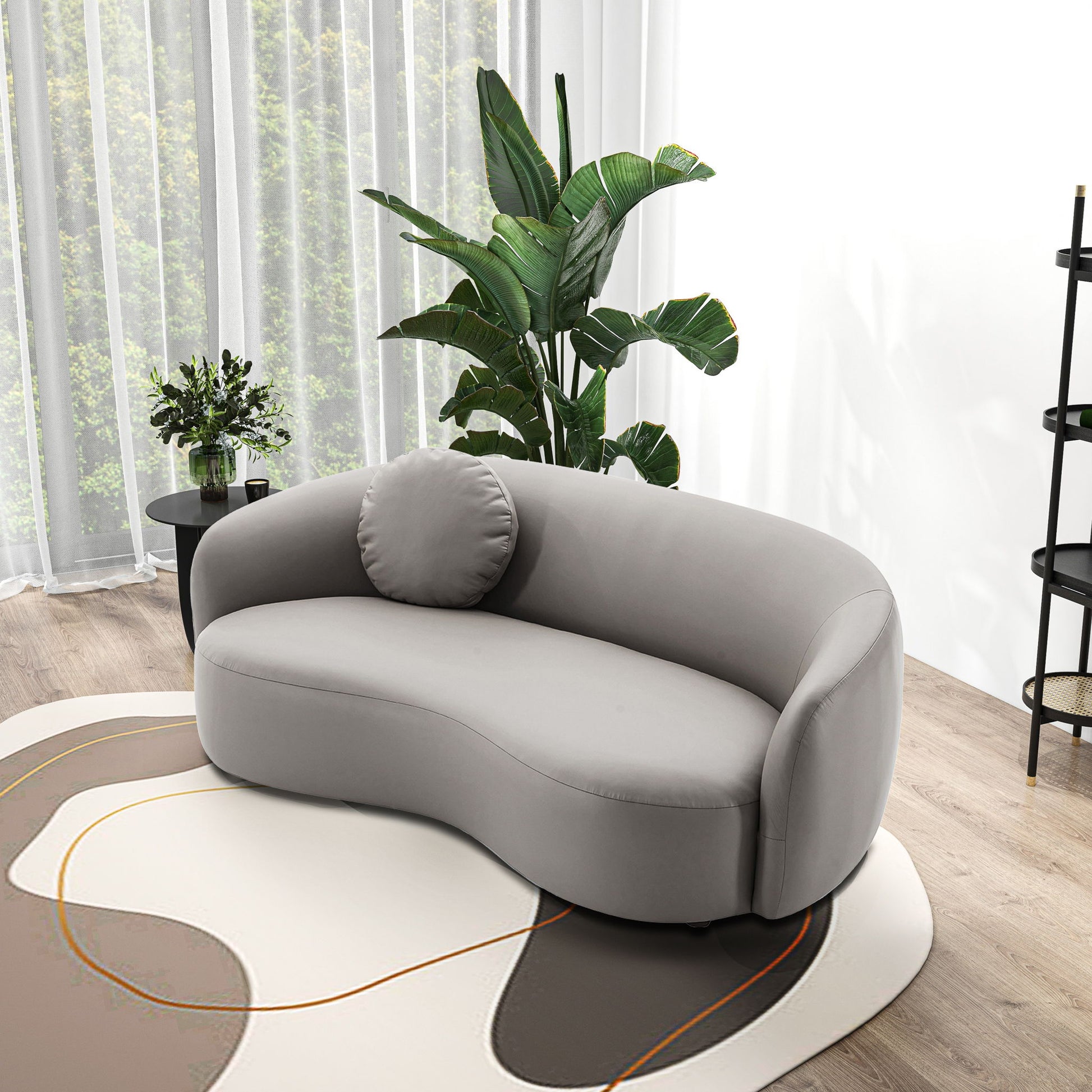 A modern living room featuring the Porter - Comfortable Sofa in gray, a round cushion, a patterned rug, green potted plants, sheer curtains, and a side table with a vase of flowers on a wooden floor.