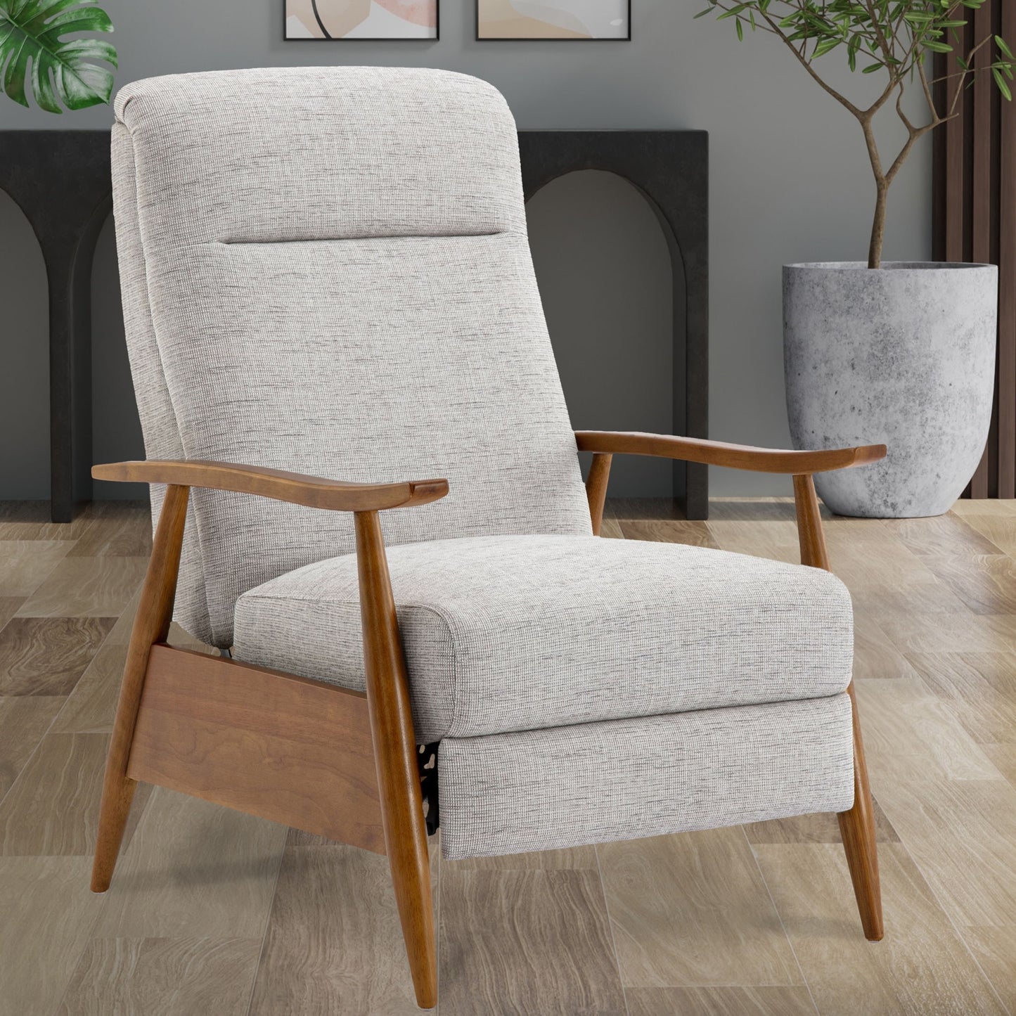 The Vaughn Push Back Recliner with solid hardwood legs and light gray upholstery sits stylishly on a wooden floor, accented by a potted plant and decorative arches in the background.