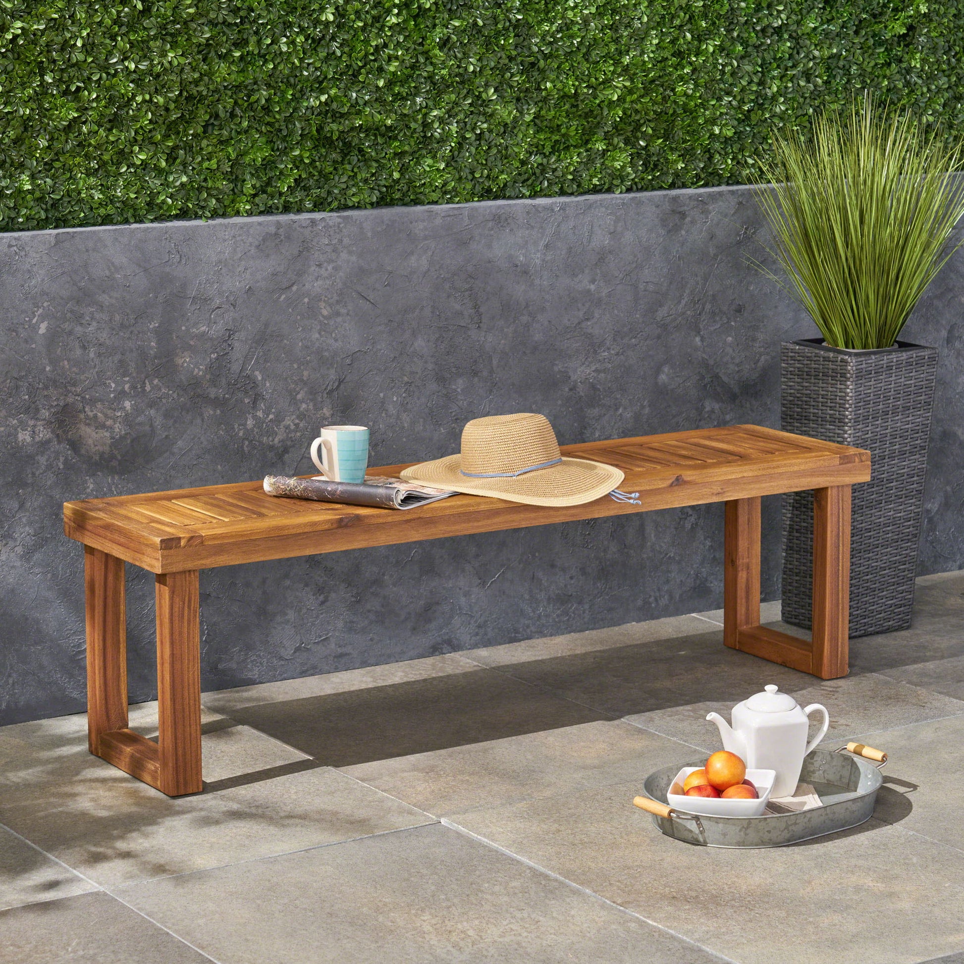 The Nestor bench sits on a stone patio against a gray wall, topped with a straw hat, mug, and newspaper. A tall potted plant stands nearby, a tray with tea and cups rests on the ground, and greenery climbs part of the wall.