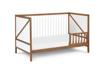 The Pixie Zen 3-in-1 Crib in Walnut is a modern wooden toddler bed with white panels and slatted sides and end, plus an open section with small vertical bars for easy access.
