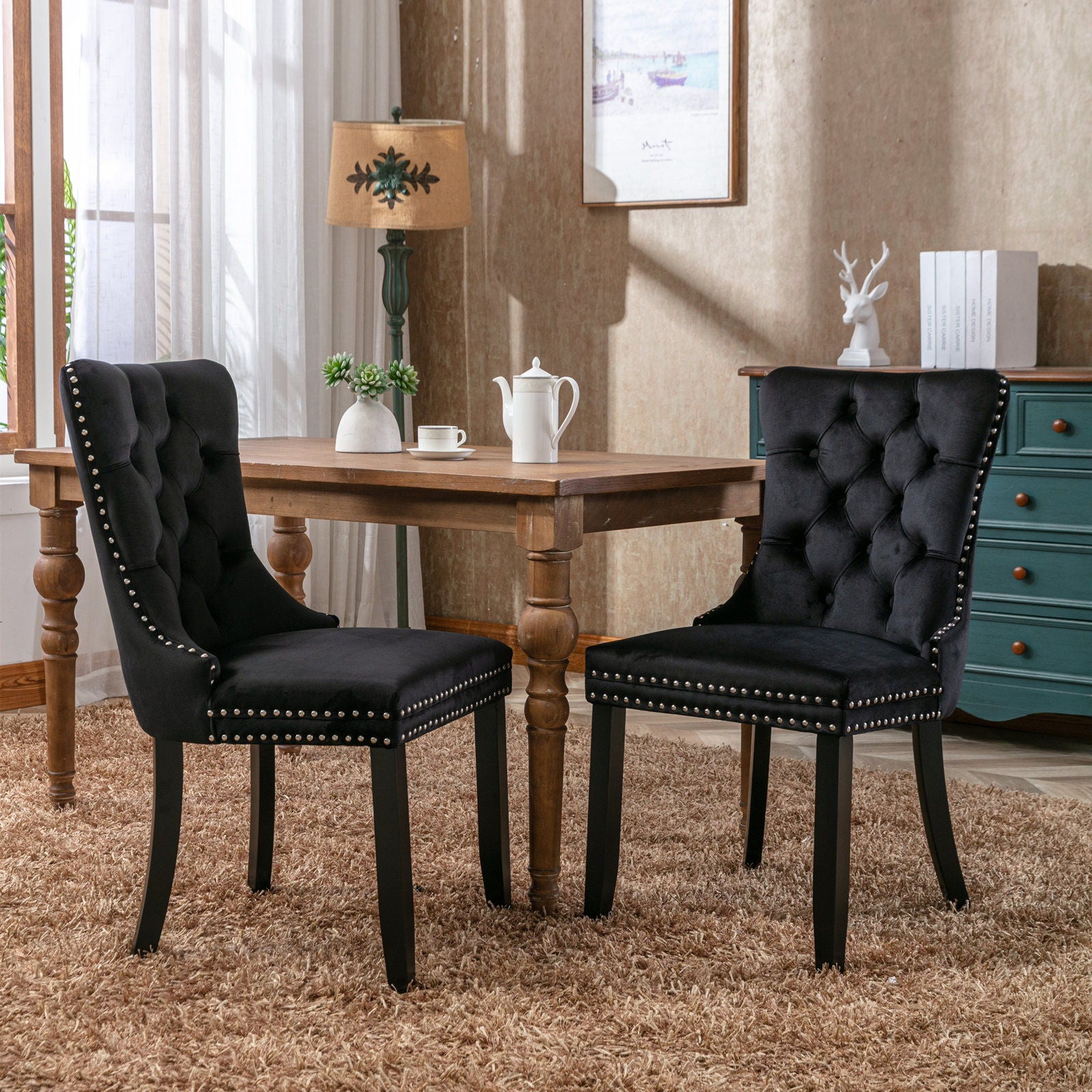 Two Nikki Modern Tufted Velvet Dining Chairs with wood legs and nailhead trim are arranged around a wooden table in a cozy room featuring beige walls, a blue dresser, a lamp, framed art, and a plush brown rug.