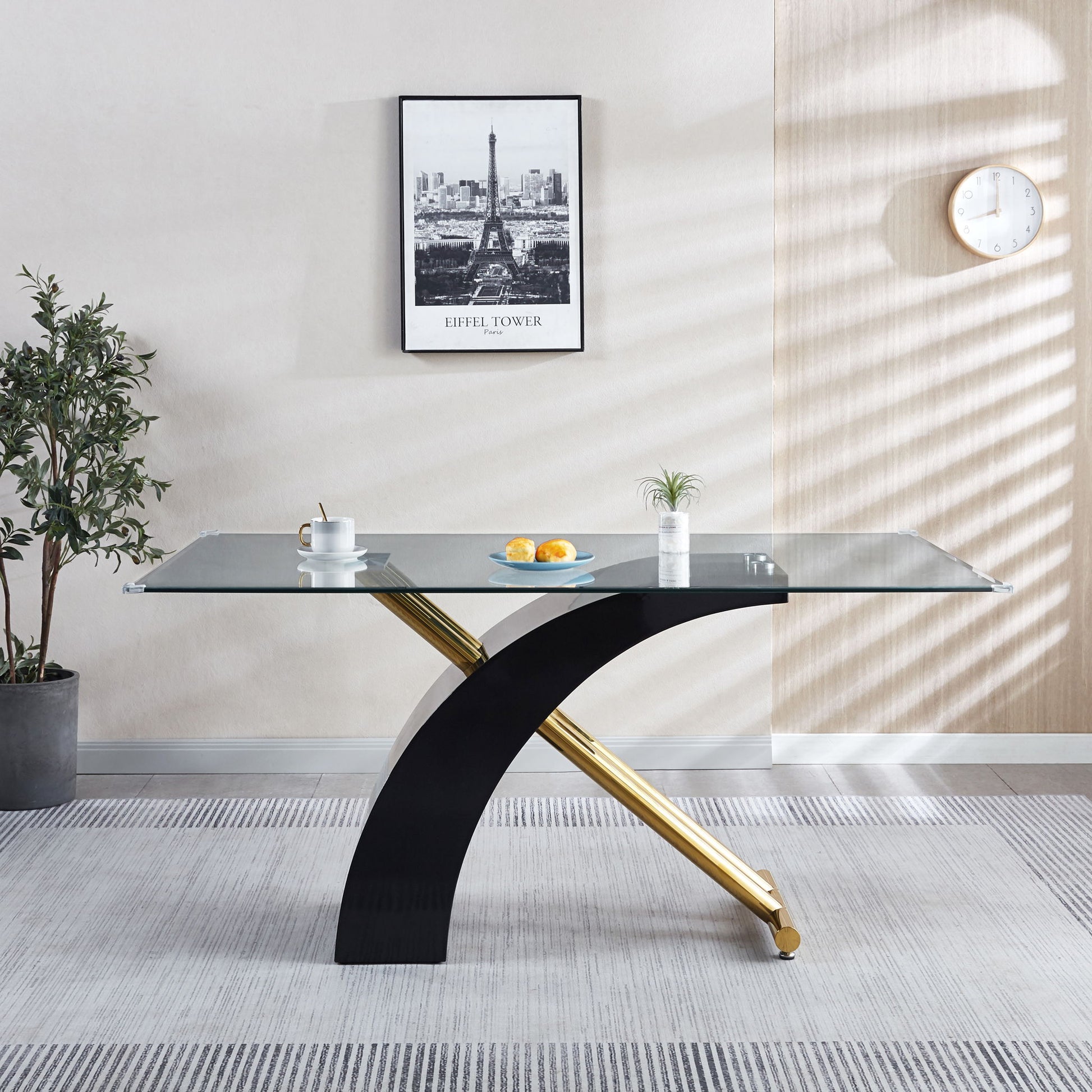 A modern dining room features the Stylish Dining Room Table with a luxury glass top and black legs, accented by a potted plant, framed Eiffel Tower photo, white clock on the wall, and two white coffee cups with lemons and a small plant.