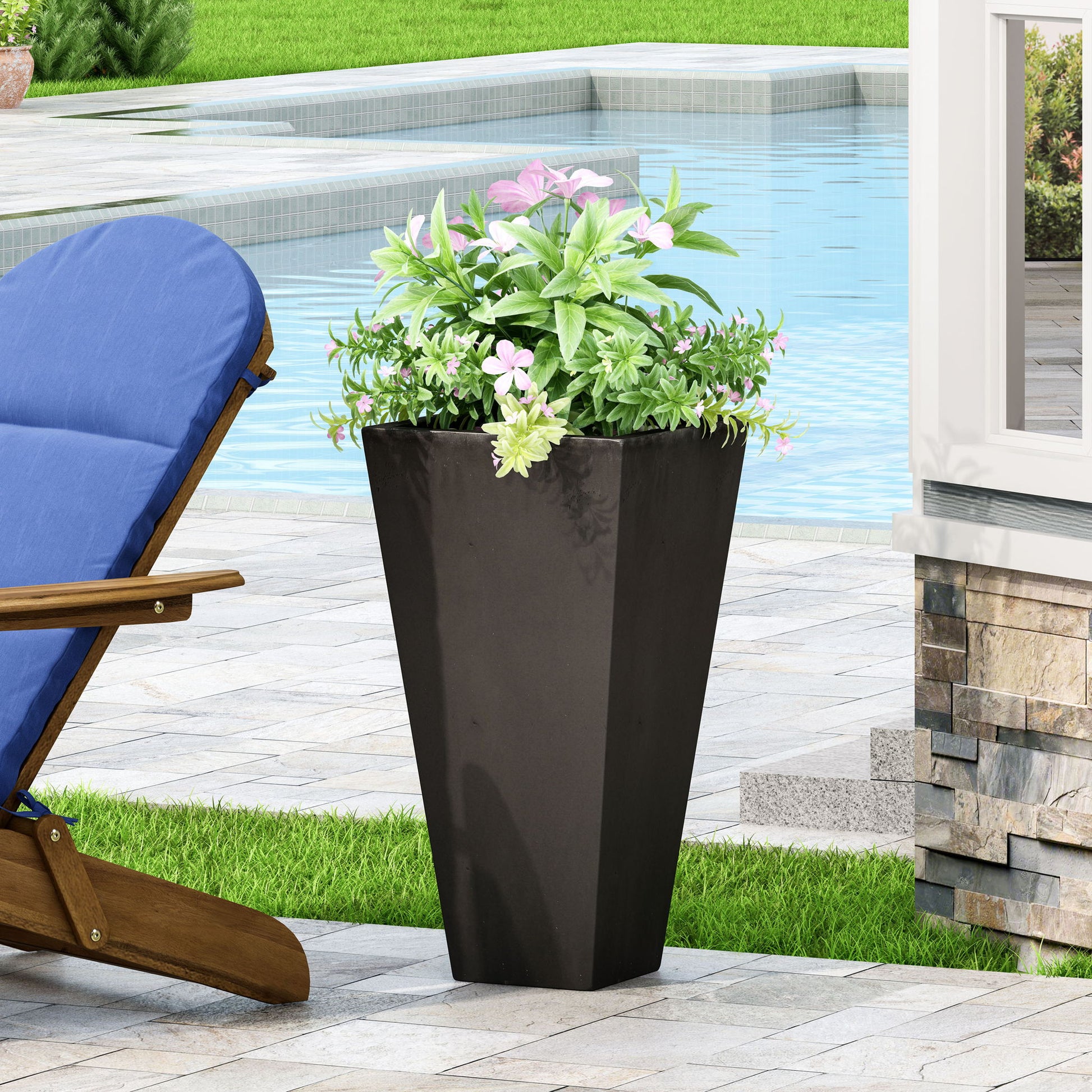 An Outdoor Modern Cast Stone Planter with lush green foliage and pink flowers sits on a patio near a pool; behind it are a blue cushioned wooden lounge chair and a stone wall.