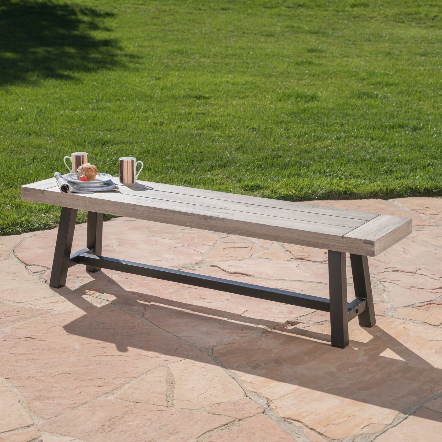 The Tranquilizer Outdoor Acacia Wood Bench with Slat Panel Design, featuring black metal legs, sits on a stone patio with a tray of striped mugs, a fork, and a pastry. Grassy lawn and tree shadows appear in the background.
