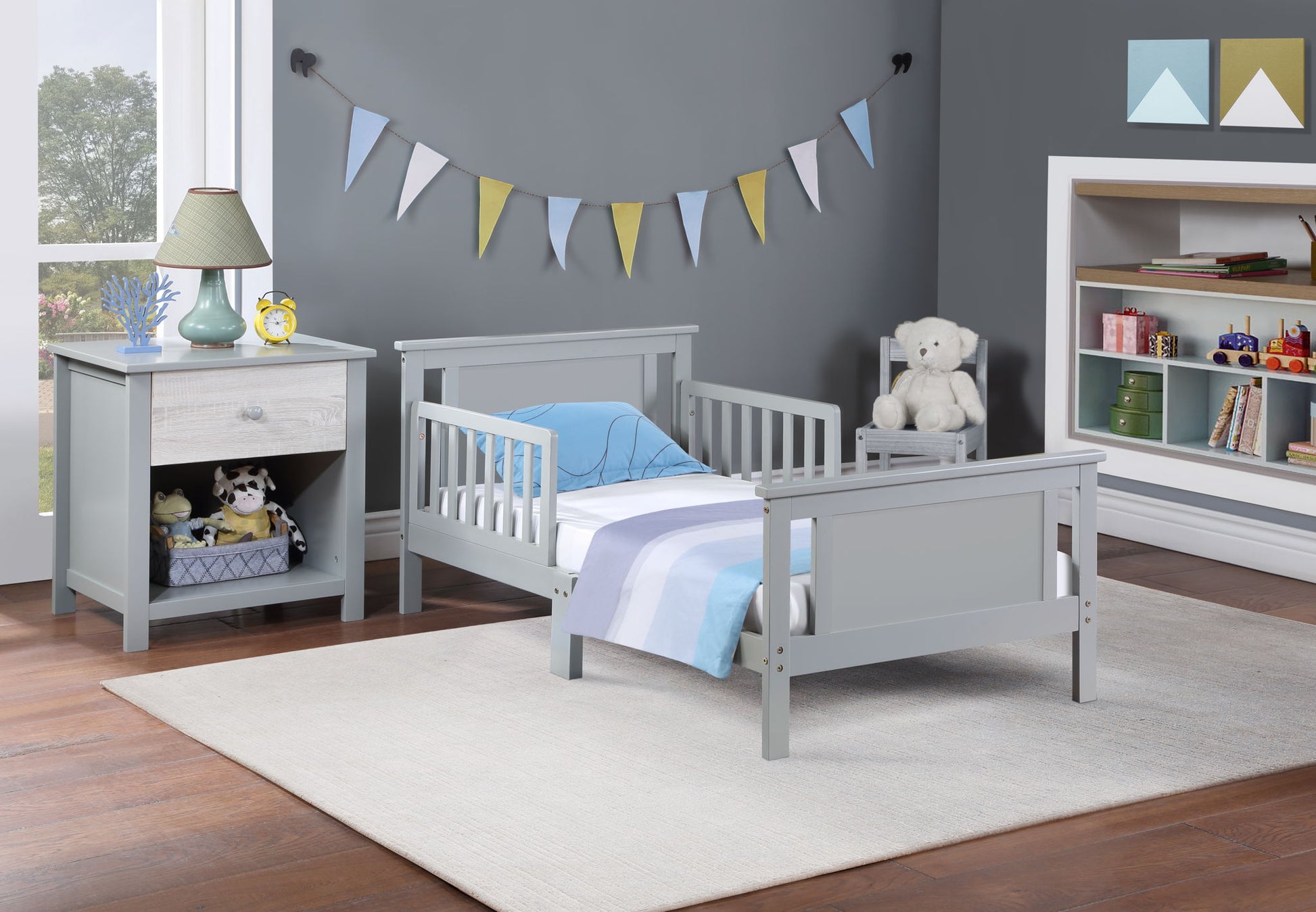 A toddler bedroom featuring the Connelly Reversible Panel Toddler Bed in gray, blue and white bedding, a gray nightstand with lamp and stuffed animals, a plush bear on a chair, wall bunting, shelves with toys and books on a gray wall.